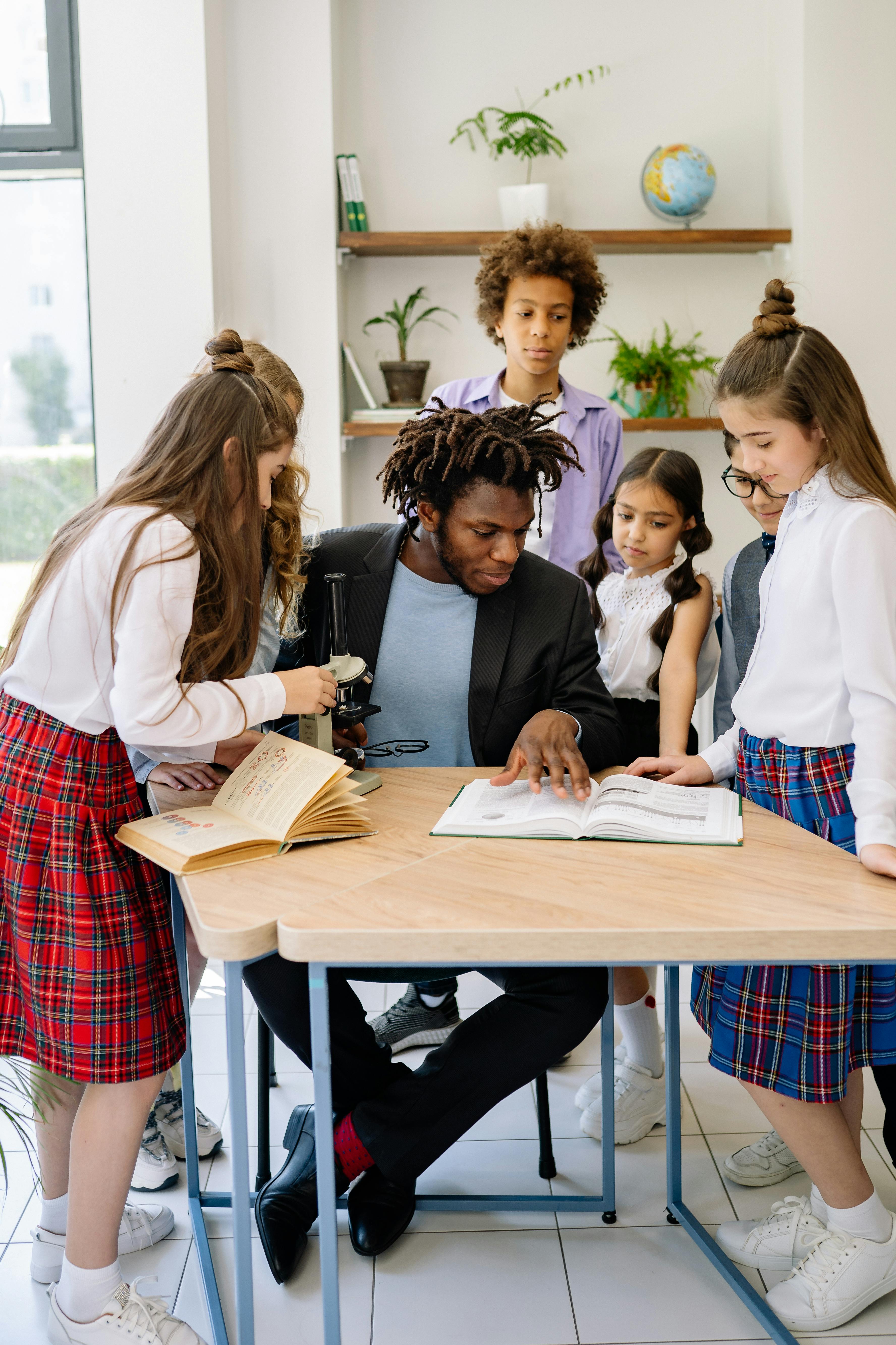 a man in black suit talking to his students while reading a book on a wooden table