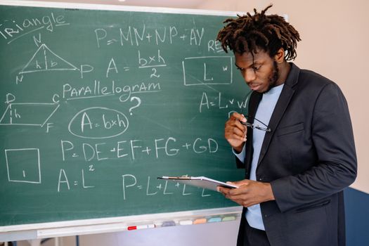 Educator in suit jacket solving math equations on blackboard, holding clipboard.