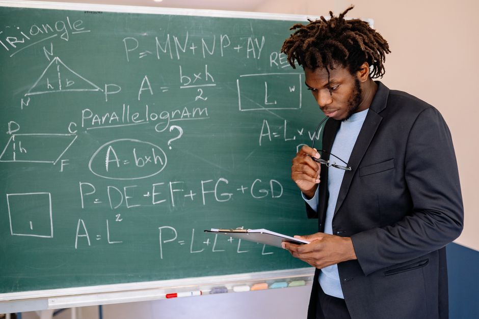 Educator in suit jacket solving math equations on blackboard, holding clipboard. Educator in suit jacket solving math equations on blackboard, holding clipboard.