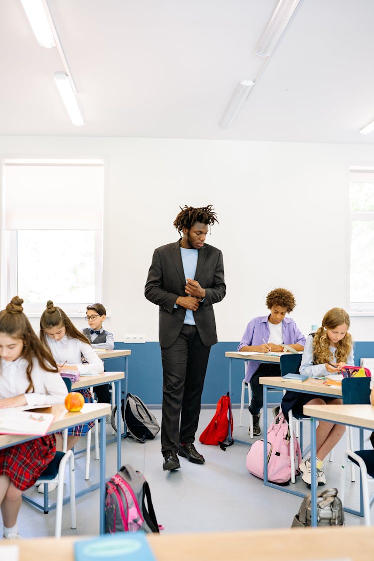 A Man Standing In A Classroom Looking At Students Sitting At Their Desks