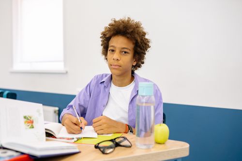 Free Young boy with afro hair writing at desk in classroom, surrounded by books and an apple. Stock Photo