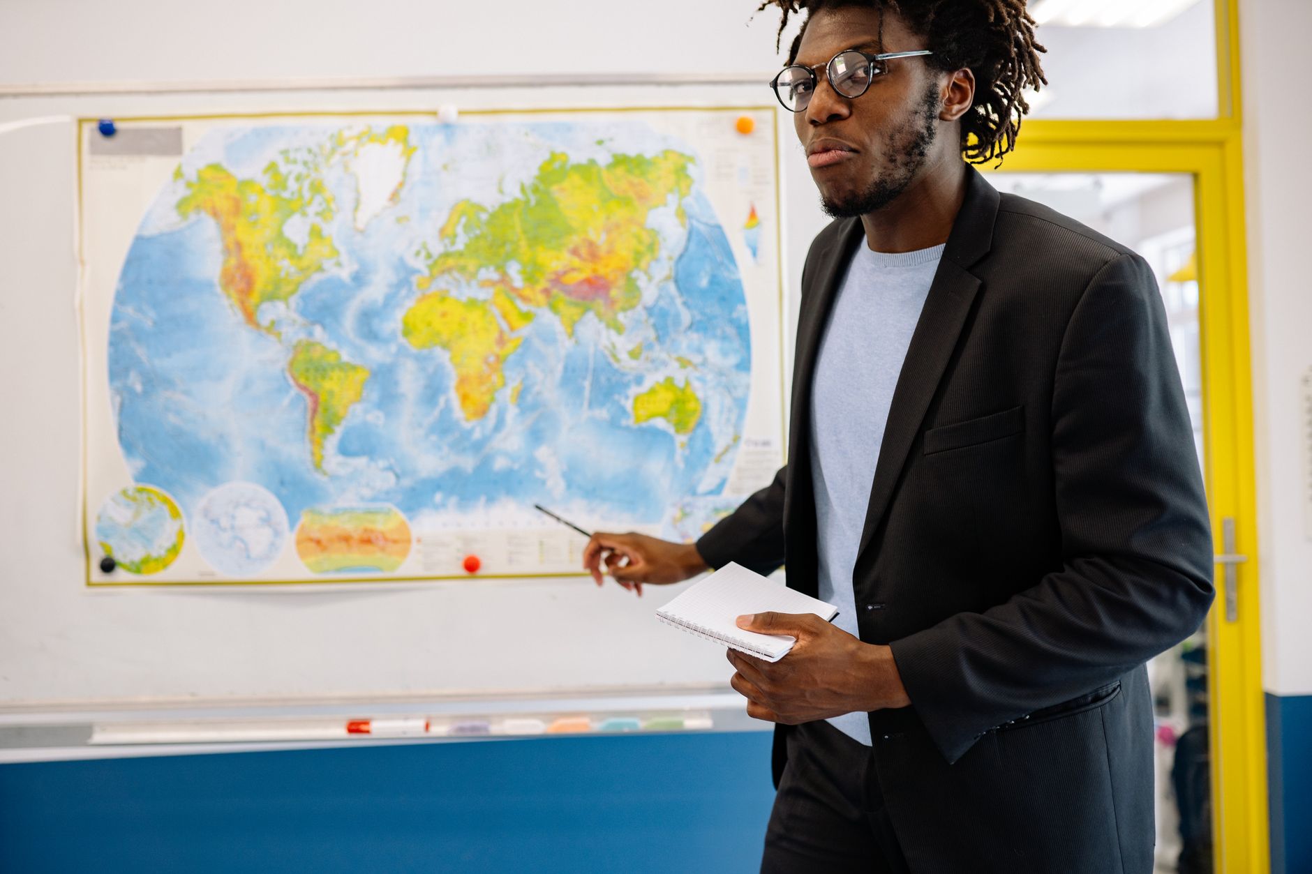 Confident teacher in a classroom explaining geography concepts using a world map.