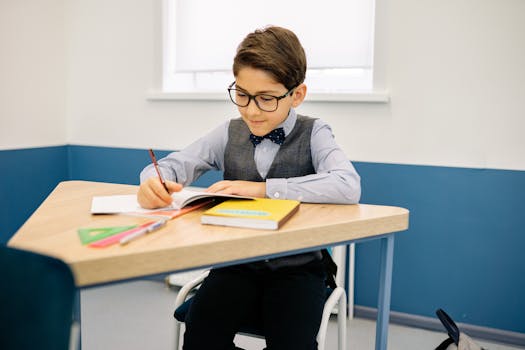 A young boy with glasses sits at a desk, writing in a notebook in a classroom.