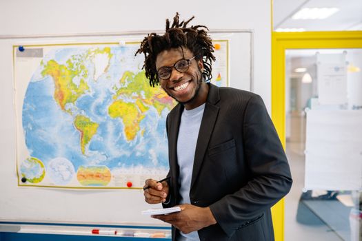 Energetic teacher smiling in front of a world map, holding a notepad, wearing eyeglasses.