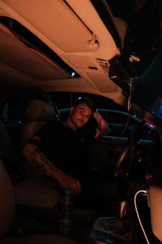 A young man with tattoos sits inside a car at night, lit by ambient lights.