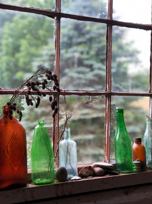 Free Colorful vintage glass bottles displayed on a rustic window sill with a scenic outdoor view. Stock Photo