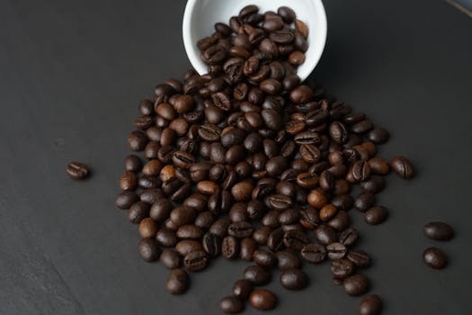 A detailed close-up of roasted coffee beans spilling out of a white cup onto a dark surface.