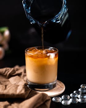 Close-up of iced coffee being poured into a glass with a dark background and a rustic setup.