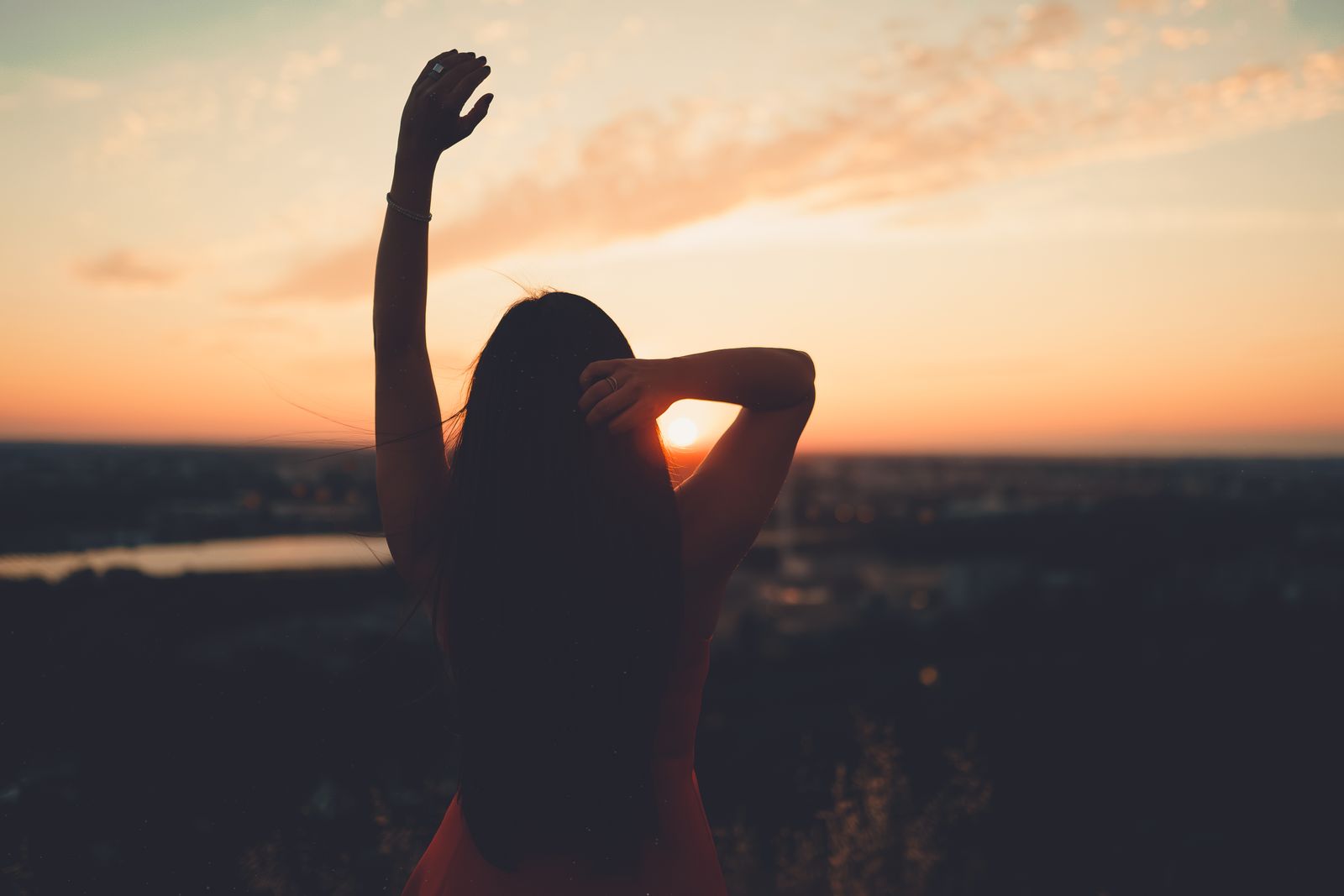 Back view of unrecognizable female raising arm and touching hair while admiring sundown on horizon