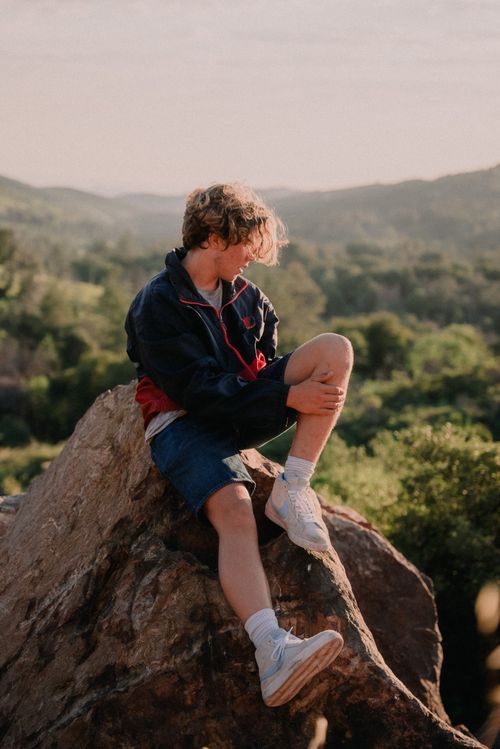Free A young man in casual attire sitting on a boulder, enjoying the tranquil outdoor scenery. Stock Photo