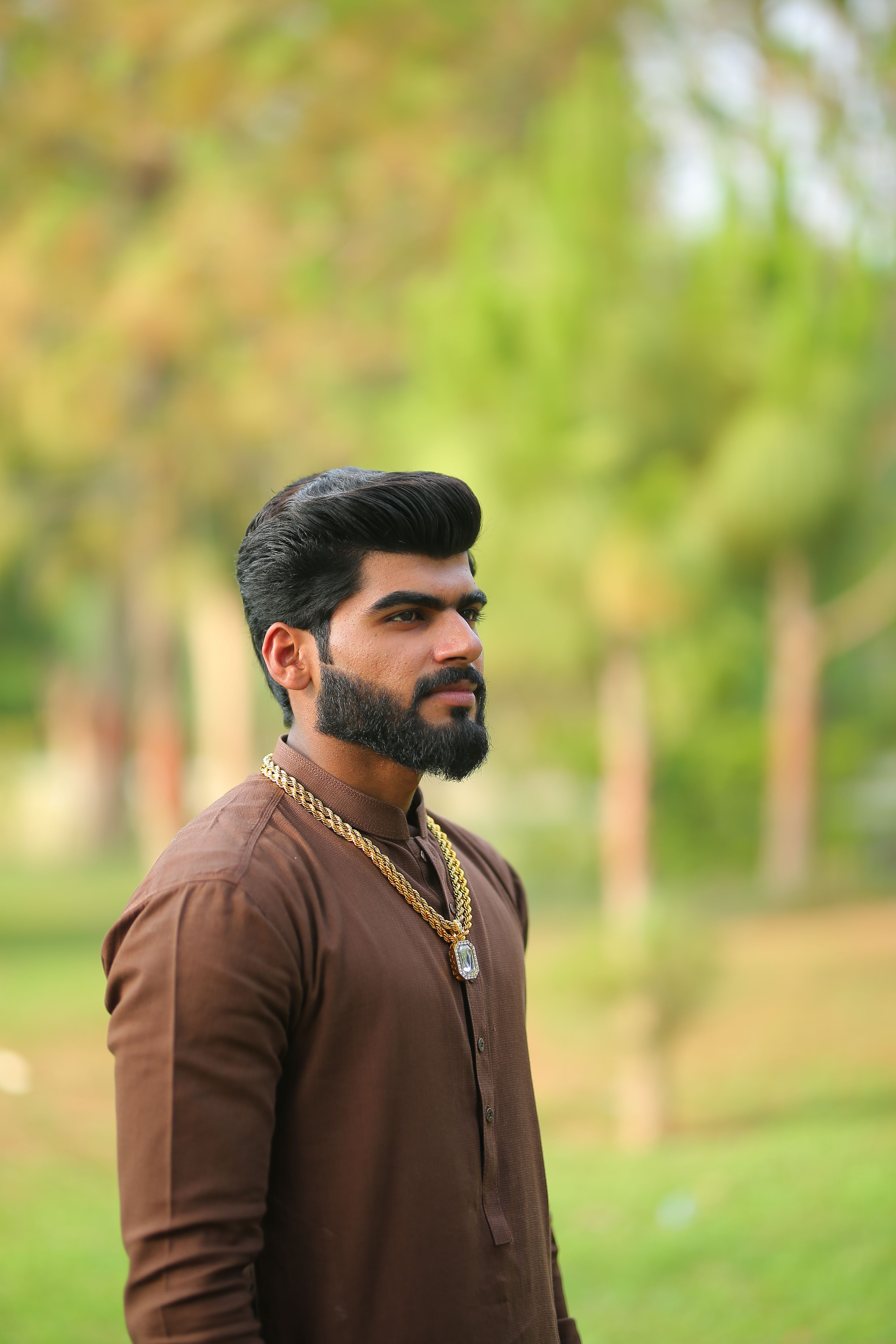 A handsome man with a beard and gold necklace standing outdoors with a blurred background.