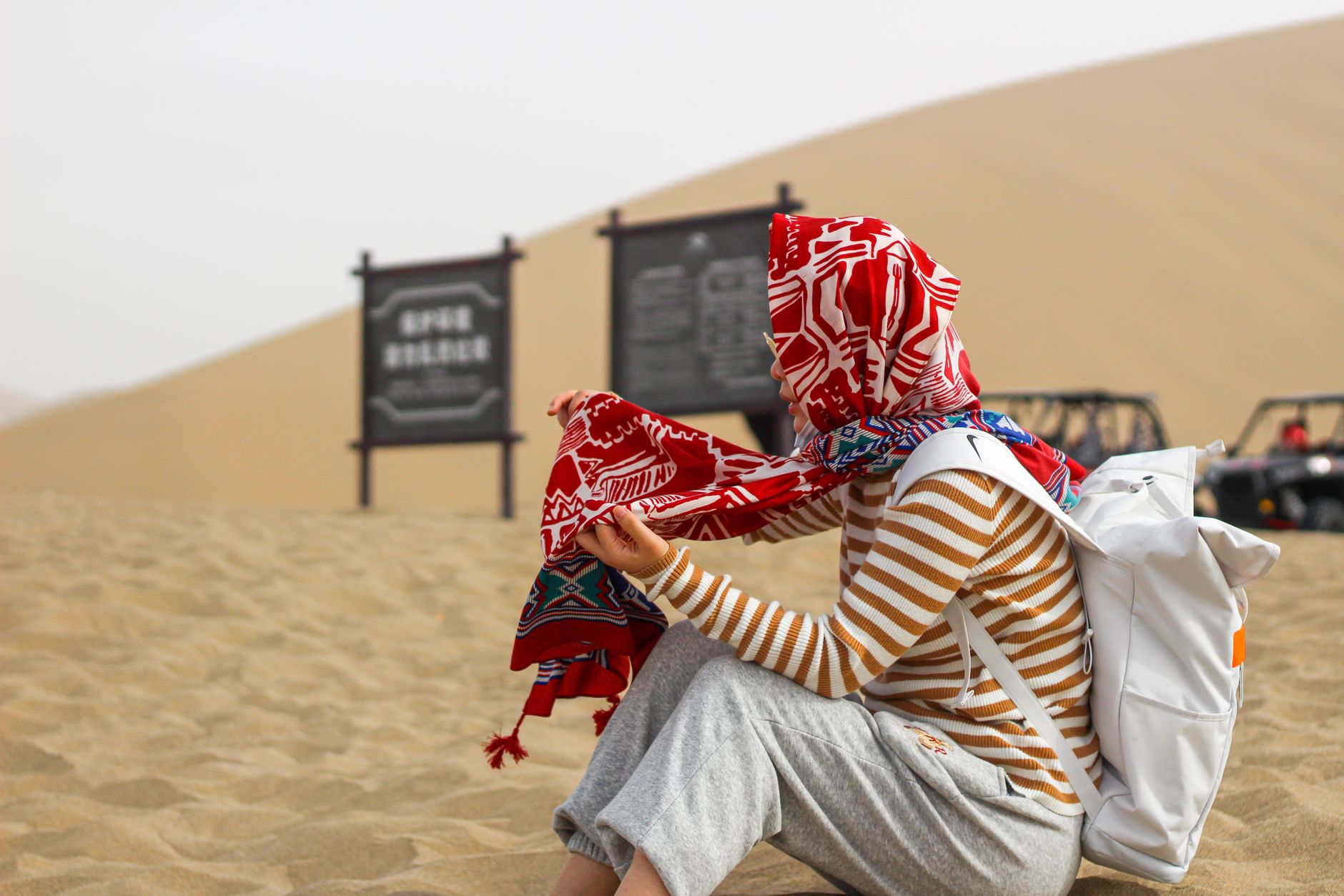 Person with headscarf sitting on sand with backpack in a desert setting.
