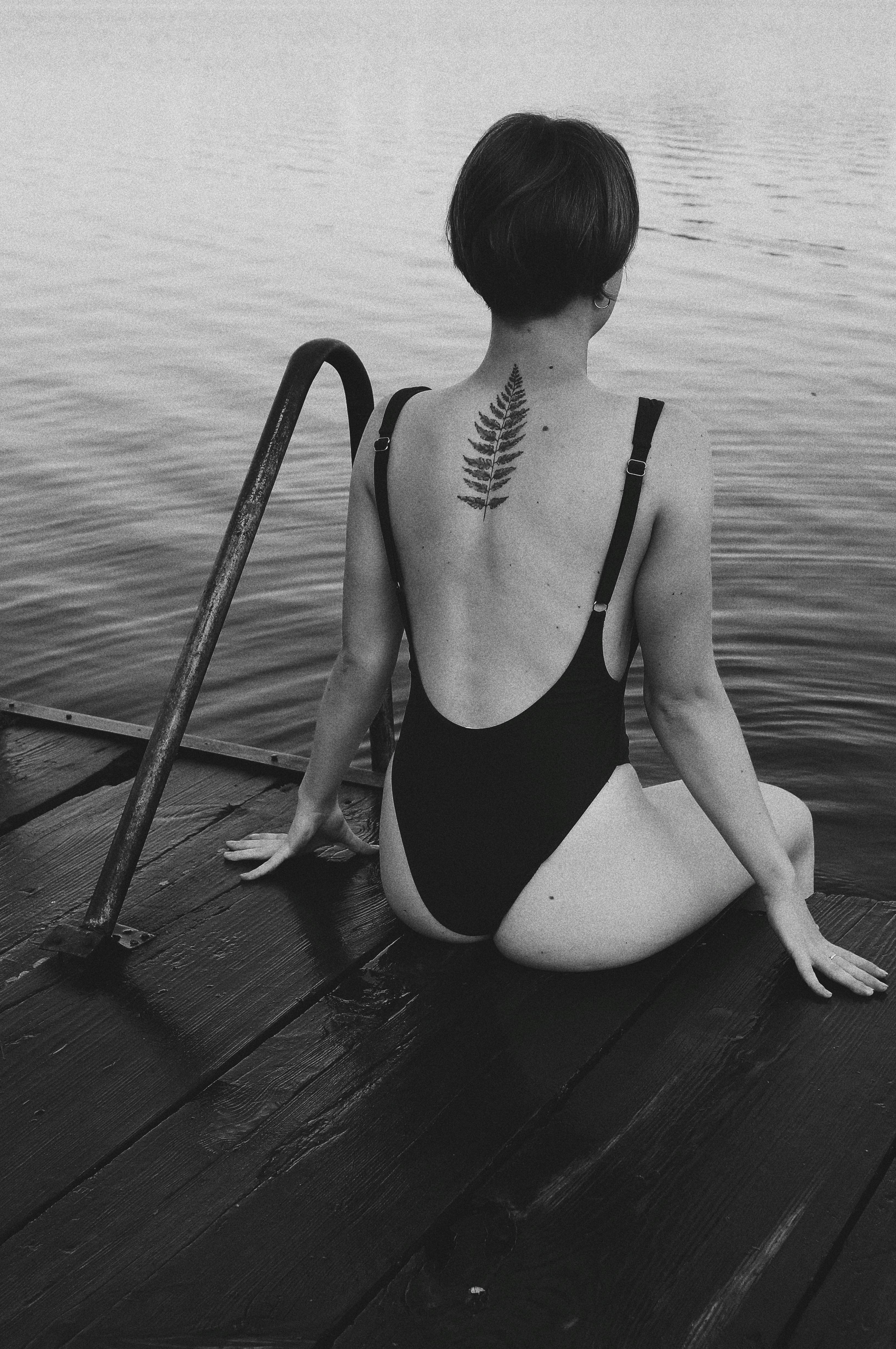 A woman in a swimsuit with a back tattoo sits on a pier, gazing at a calm lake.