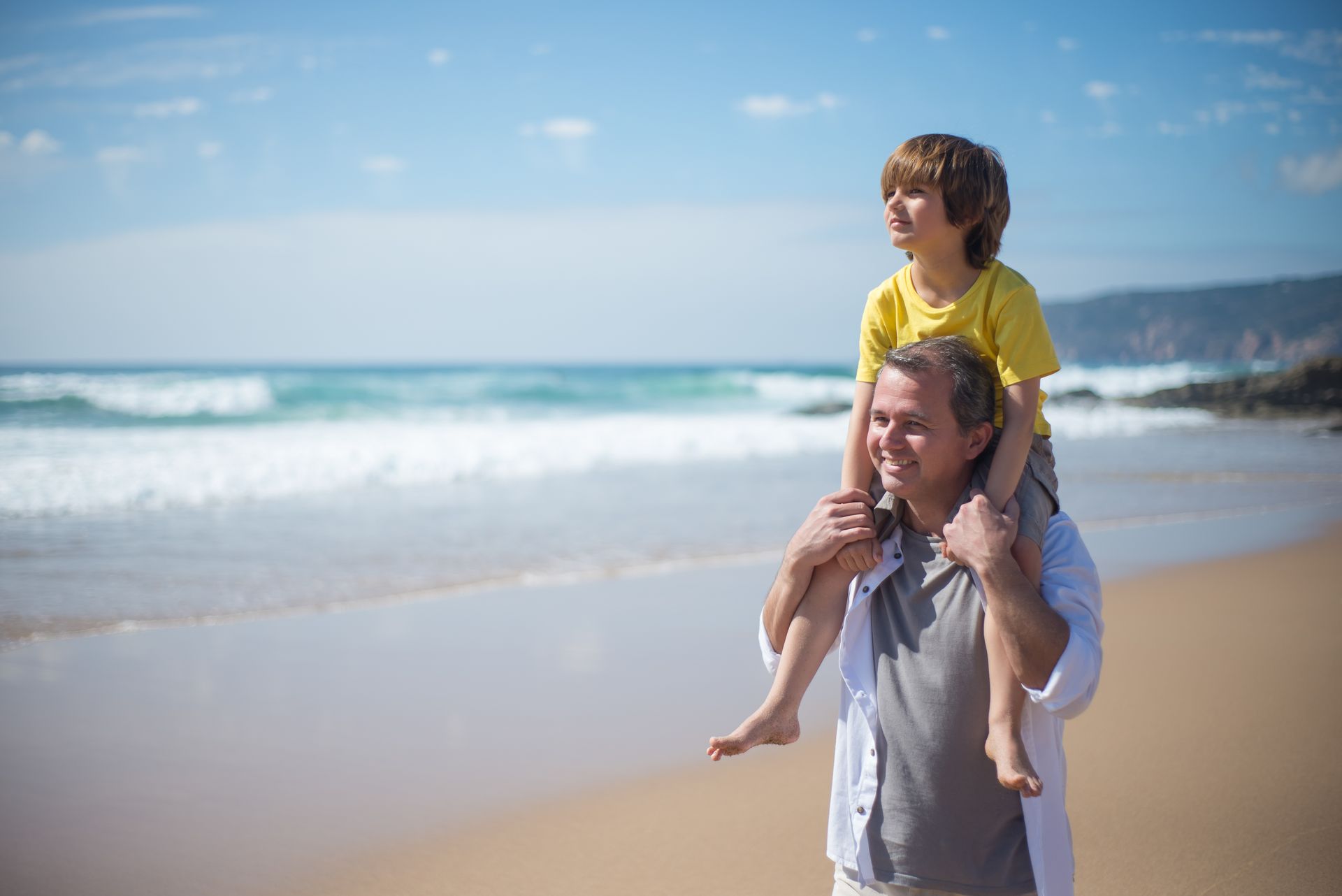Father carrying son on shoulders during a sunny beach day in Portugal.