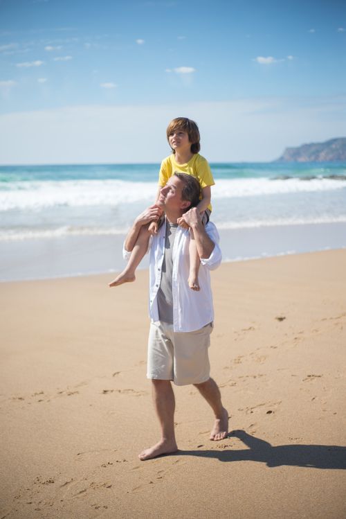 Free A father carrying his son on shoulders along a sunny Portuguese beach, capturing a moment of joy and togetherness. Stock Photo