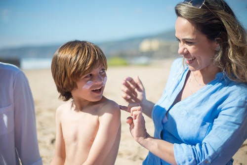 Woman in Blue Long Sleeve Shirt Putting Sunscreen Cream on Boy's Back