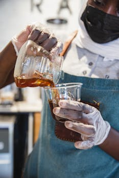 A barista wearing gloves pours coffee into a cup, showcasing a hygienic cafe setting.