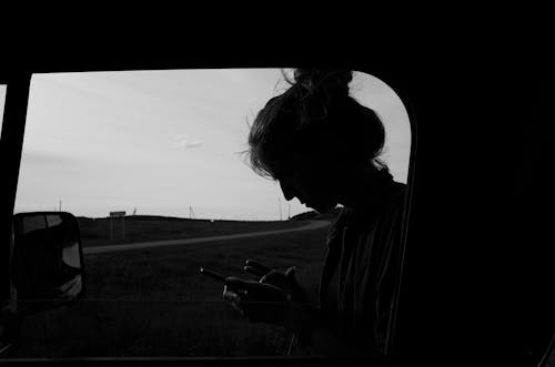 Grayscale Photo of a Person Using a Cellphone beside a Car Window