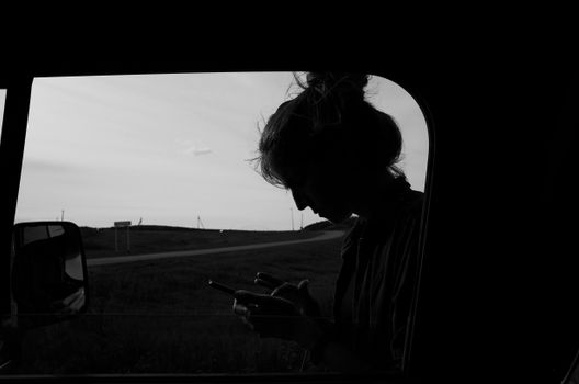 Black and white silhouette of a woman using a smartphone inside a car window.