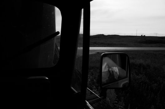 Black and white image capturing a person's hand reflected in a car side mirror against a rural backdrop.
