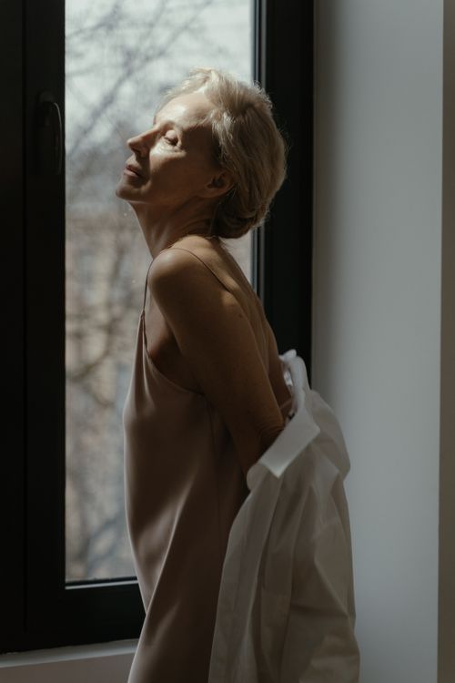 Free Senior woman in a nightdress by a window, basking in soft natural light. A serene and contemplative moment indoors. Stock Photo