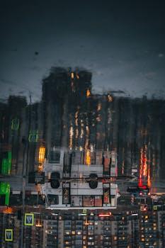 A bus and illuminated city buildings reflected in a puddle at night, creating a moody urban scene.