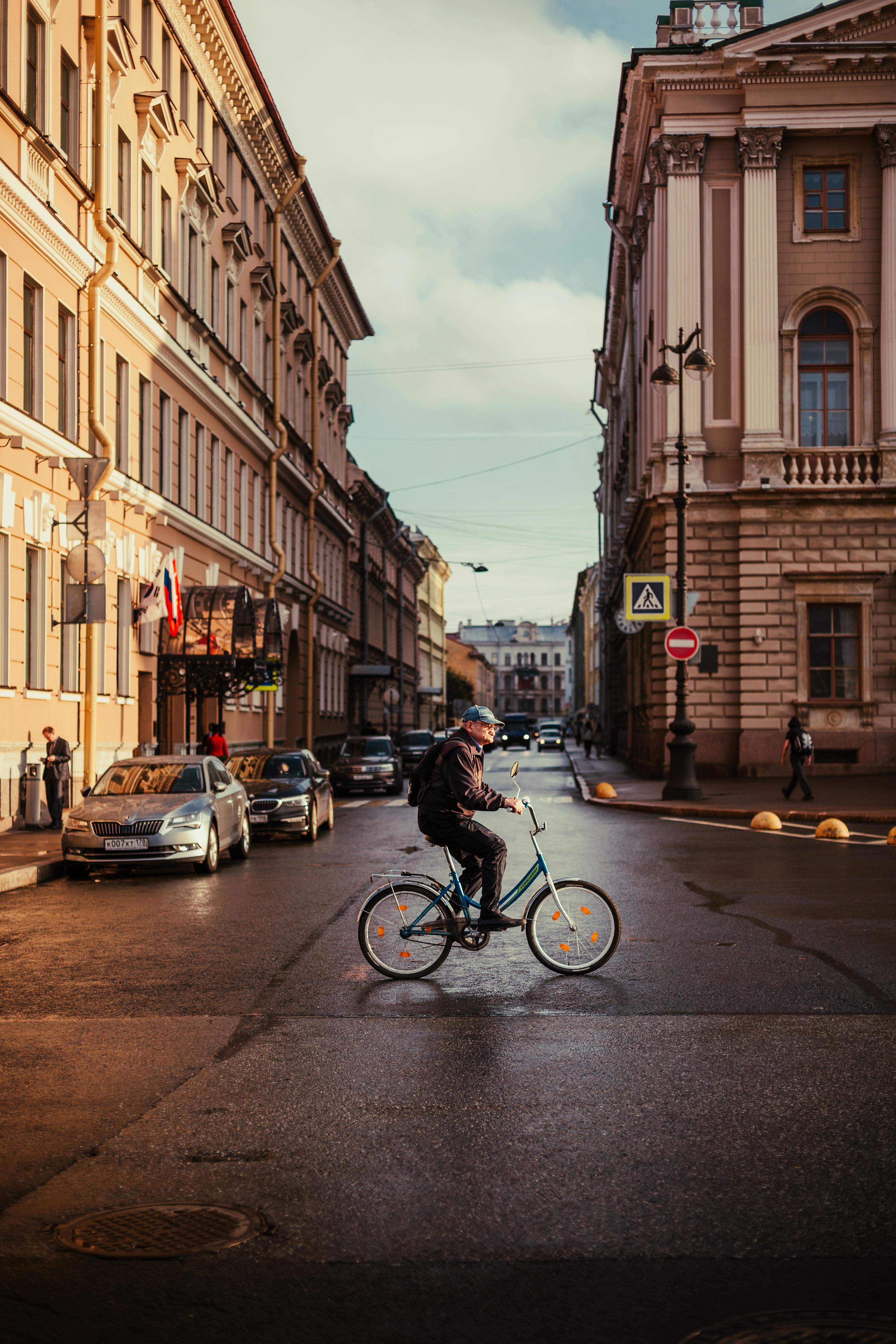 Man cycling across a European city street surrounded by classic architecture and parked cars.