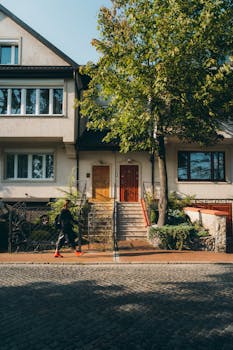 Urban street view featuring a modern residential facade, lush greenery, and a person walking on the sidewalk.
