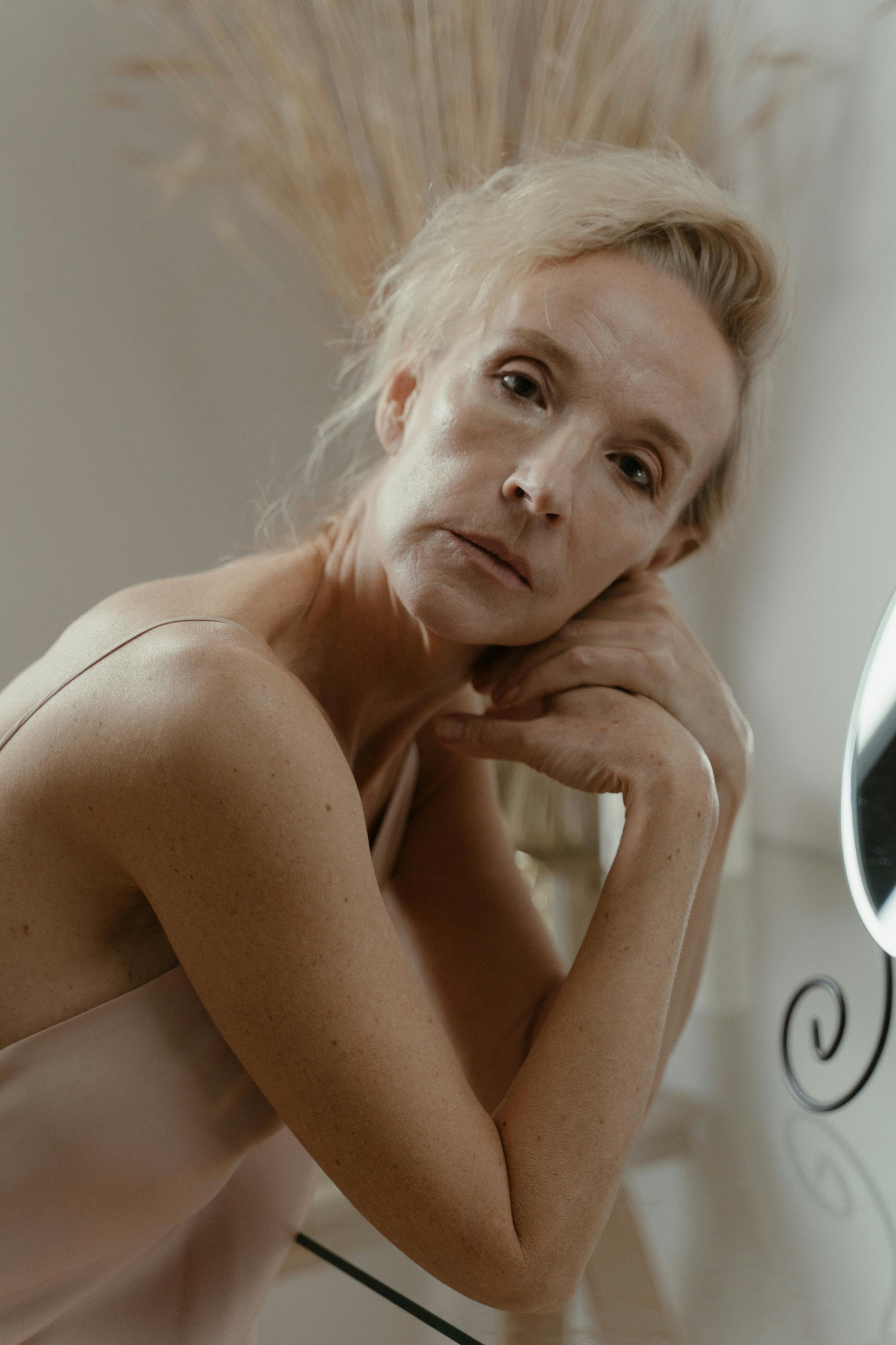 Thoughtful portrait of a senior woman sitting at a reflective glass surface indoors.