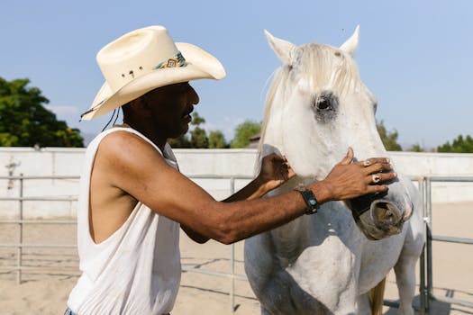 African American cowboy wearing hat bonding with a white horse outdoors.