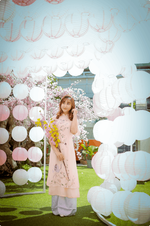 Young Woman in Pink Dress Holding a Flower and Standing on Hanging White Lanterns