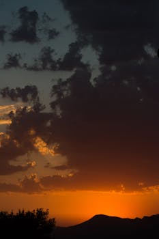 A dramatic sunset with vibrant colors and cloud formations over a silhouetted mountain scene.