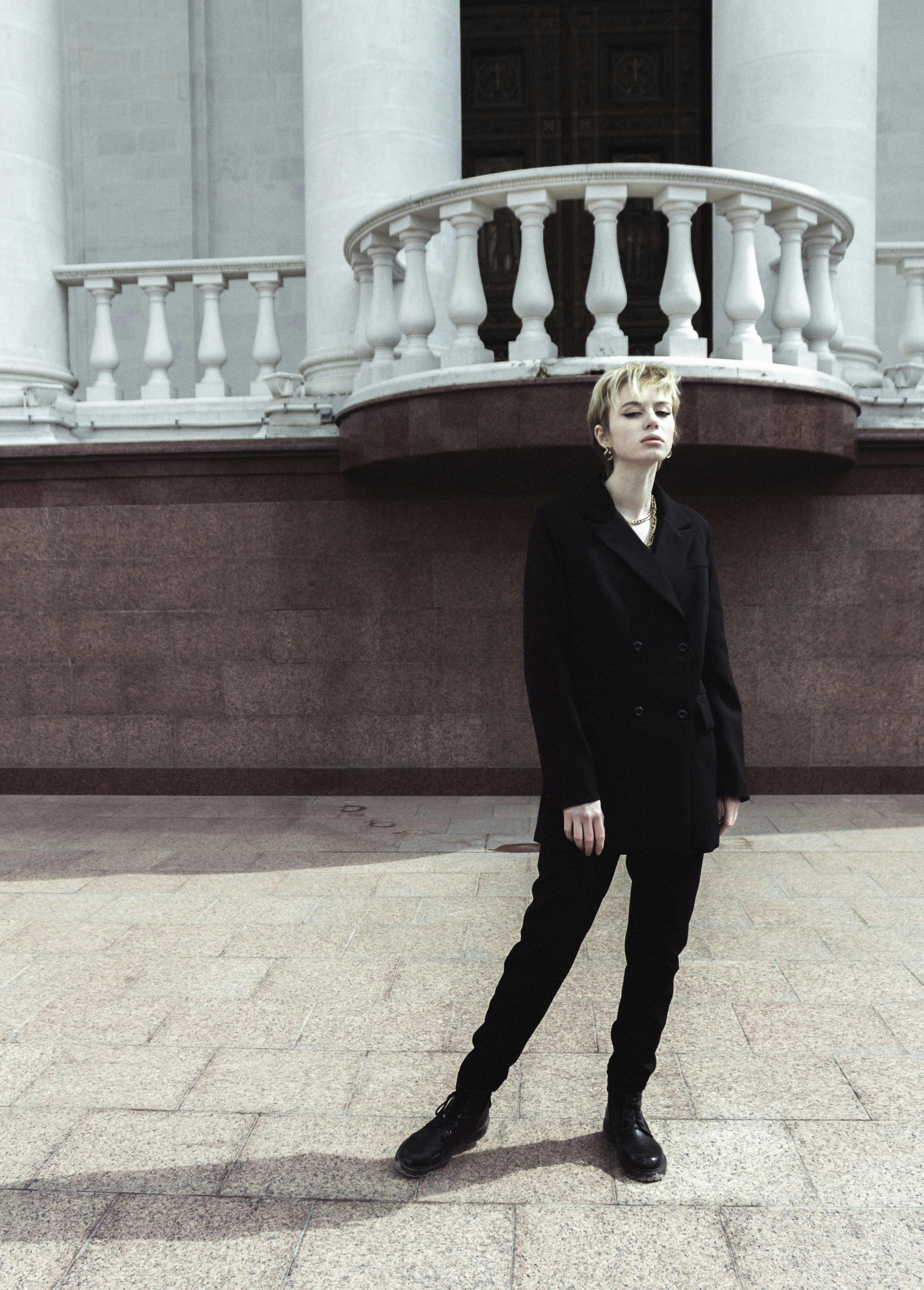 Fashion-forward individual wearing black clothes poses confidently against classical architecture.
