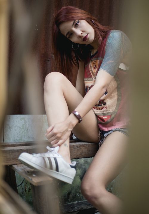 Free Stylish young woman with red hair and tattoos sitting on a bench outdoors, wearing a casual outfit. Stock Photo