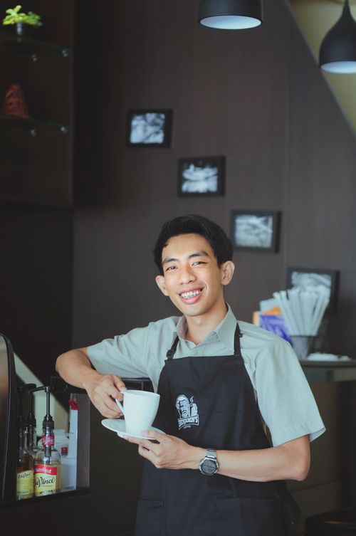 Free Cheerful barista holding a coffee cup, smiling warmly in an Indonesian cafe. Stock Photo