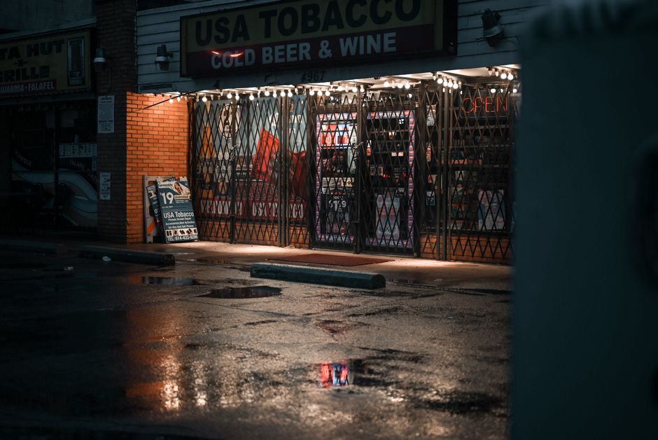 An urban storefront with gated windows at night, reflecting on wet pavement under artificial lighting.