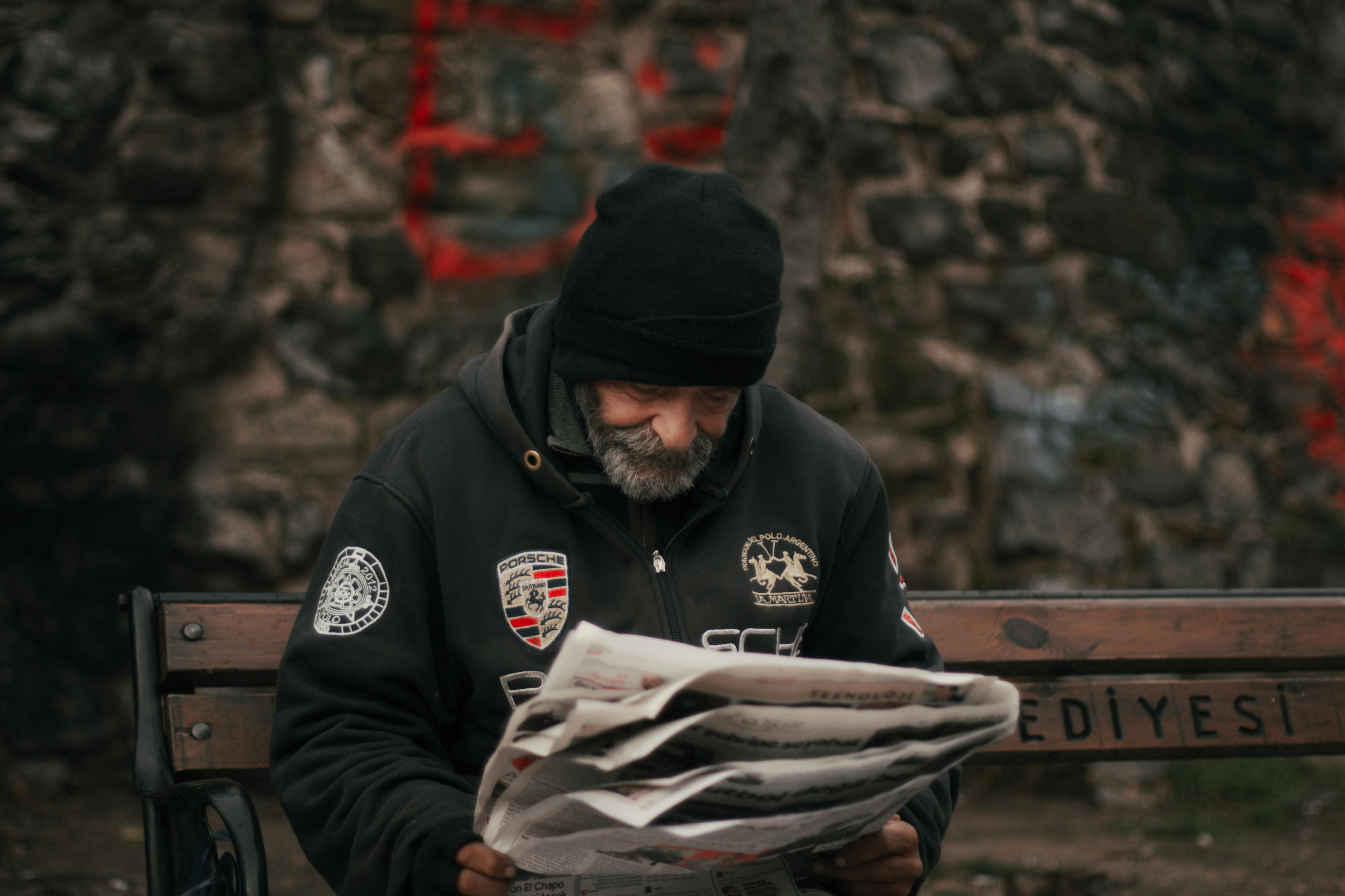 a bearded man reading newspaper while sitting on a wooden bench