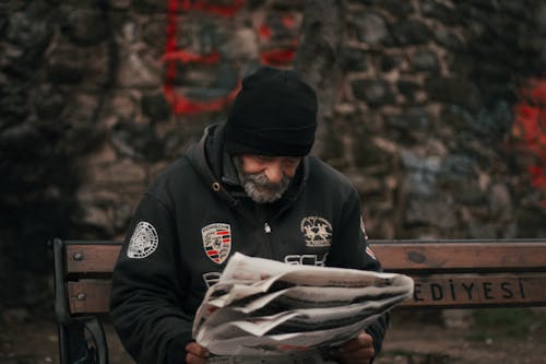 A Bearded Man Reading Newspaper while Sitting on a Wooden Bench