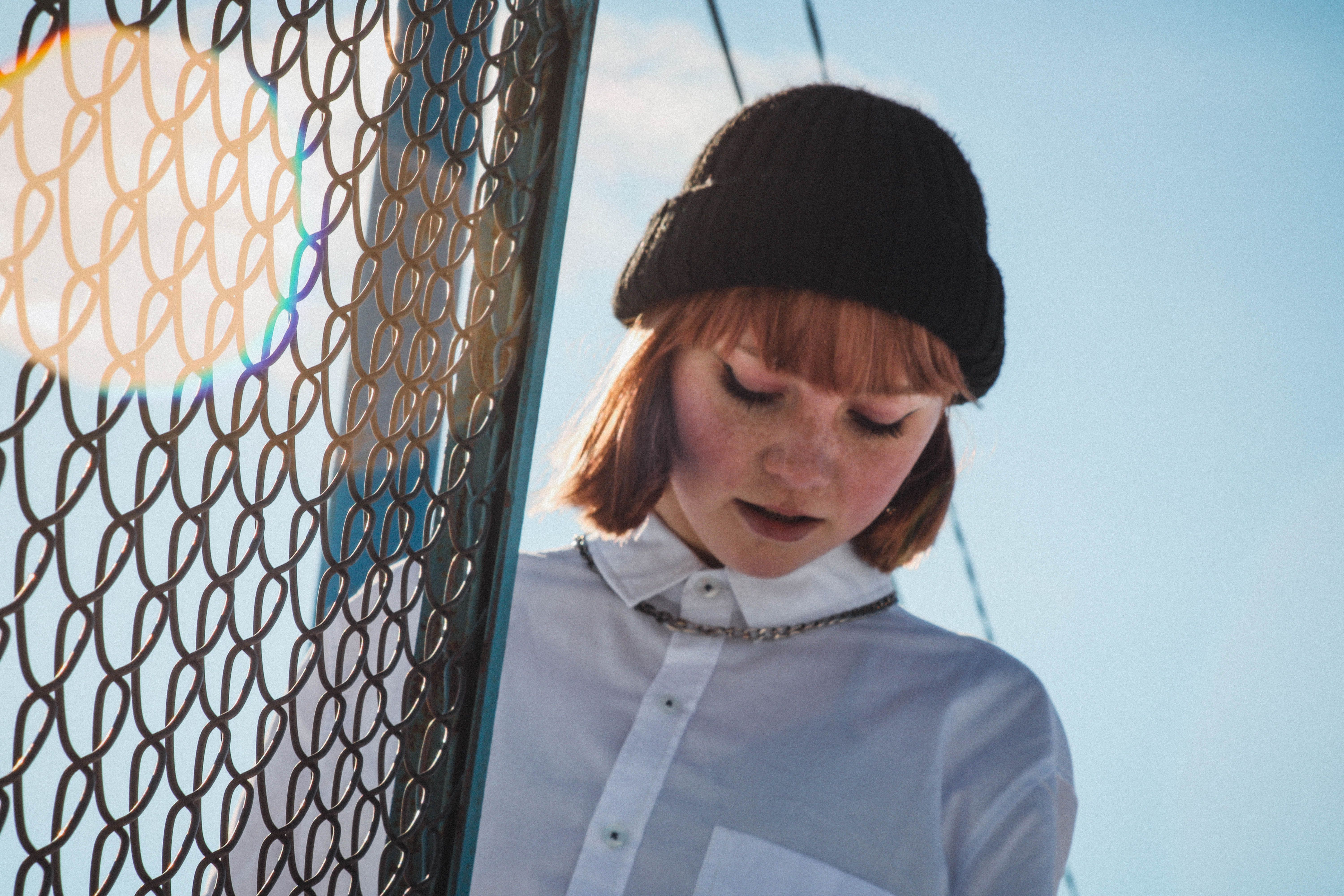 Jeune femme portant un bonnet tricoté noir à revers, chemise blanche, devant un grillage