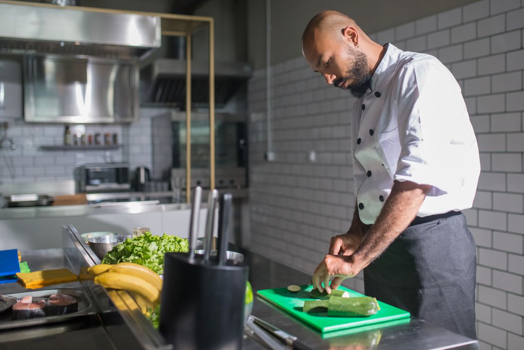 10 Essential Knife Skills Every Home Cook Must Learn 1 A professional chef in a white jacket uses the claw grip to slice fresh vegetables on a green cutting board in a modern kitchen.