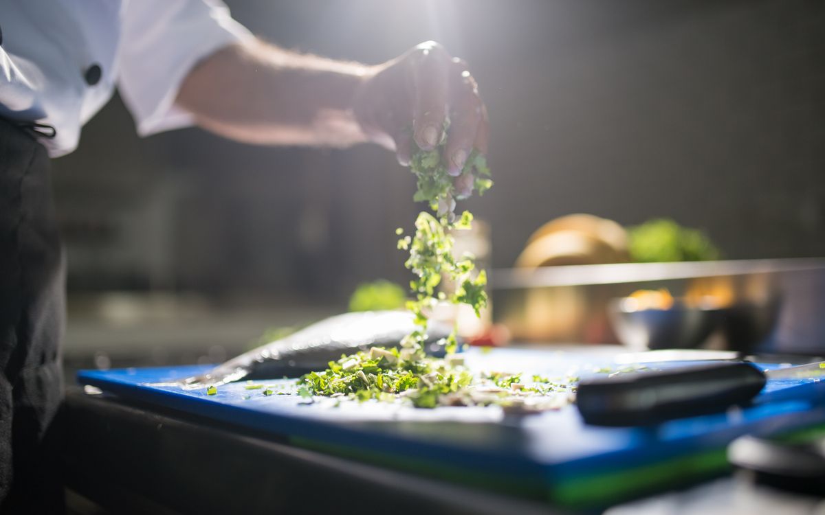 Chef's hand preparing chopped green vegetables on a cutting board in a kitchen.