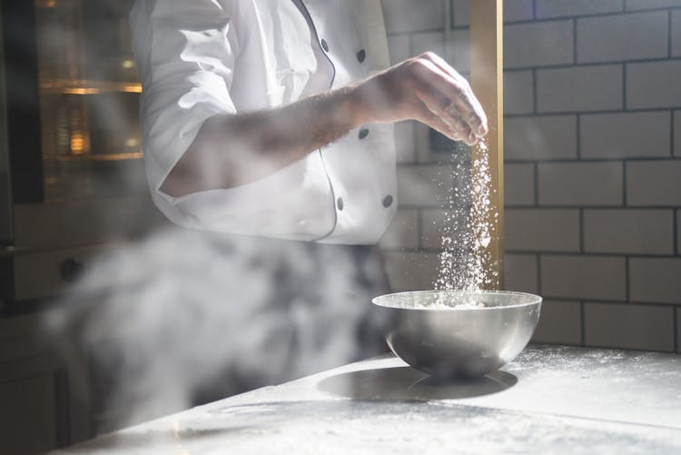 A Person Sprinkling Flour On A Steel Bowl