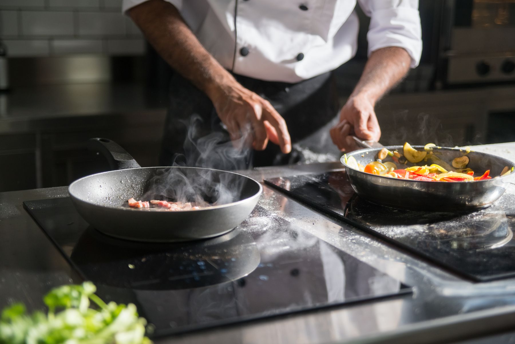 Chef in whites cooking sizzling meat and colorful vegetables in two pans on an induction range