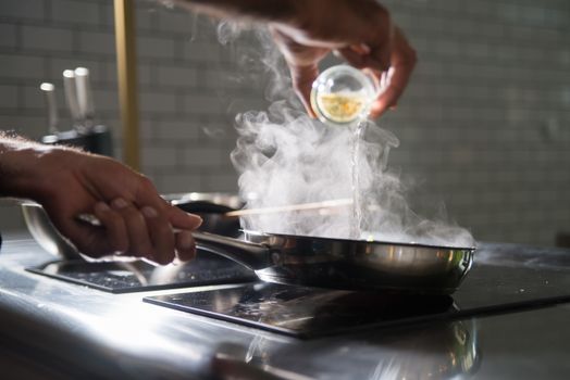 Close-up of a chef adding liquid to a steaming frying pan in a modern kitchen.