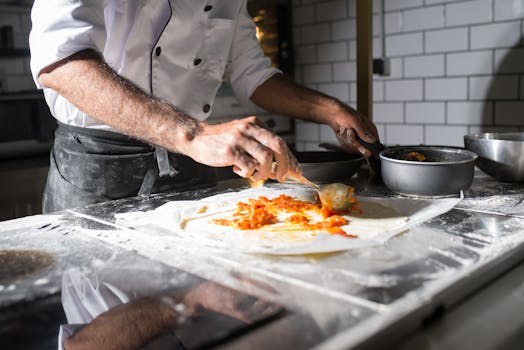 Professional chef spreading tomato sauce on dough in a commercial kitchen.