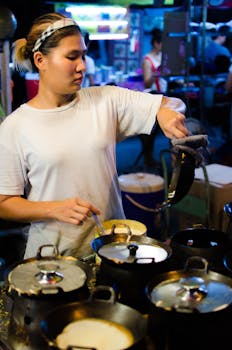 An Asian woman is focused on cooking at a bustling night market's food stall, surrounded by pots.