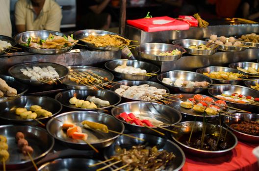 Assorted Asian street food skewers arranged on metal plates at a vibrant market.