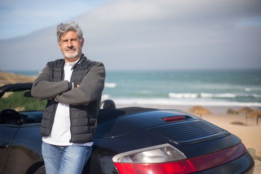 An elderly man stands confidently by his car on a scenic beach in Portugal.