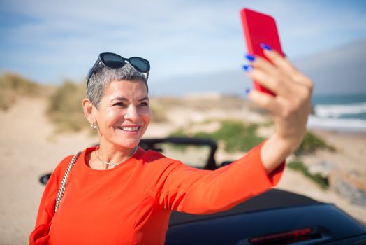 Smiling woman in red taking a selfie by the beach in Portugal with a convertible car.