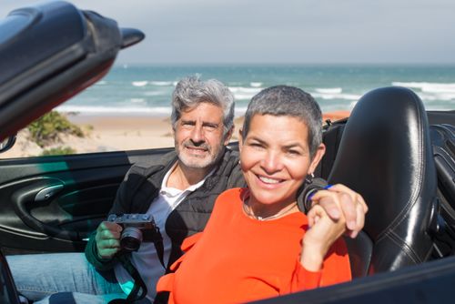 Free Happy senior couple with a camera in a convertible by the beach on a sunny day in Portugal. Stock Photo